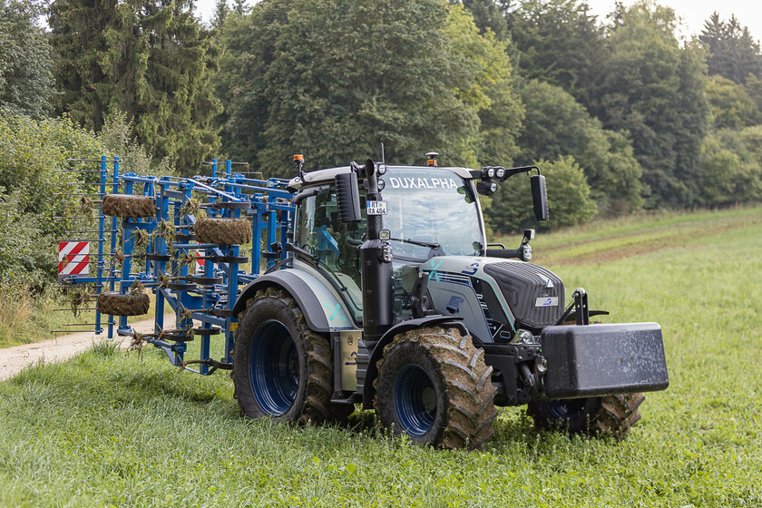 Ein silberblauer Traktor mit einem blauen Anbaugerät steht auf einer Wiese. Die Reifen und das Anbaugerät sind mit Erde bedeckt. 
