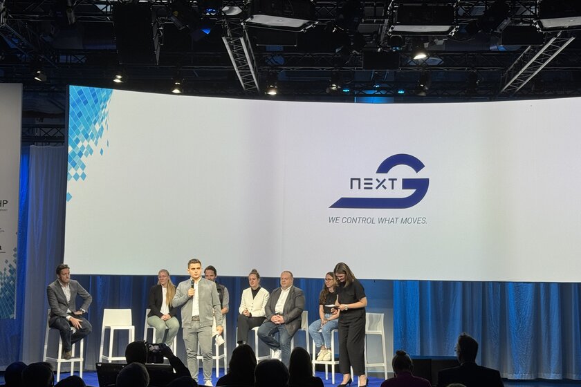 Vor einer sehr großen Leinwand auf der ein Arnold NextG Logo drauf ist, sitzen mehrere Personen auf einer Bühne. Vor ihnen steht ein junger Mann (Kevin Arnold) der in ein Mikro redet. 