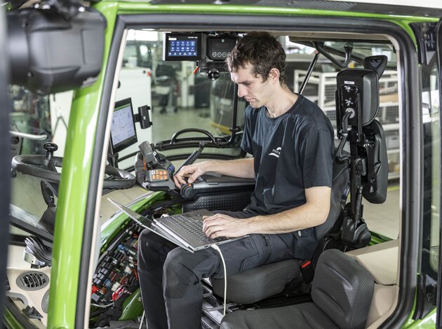 Ein Mitarbeiter der Firma Arnold NextG sitzt mit seinem Laptop, auf dem Schoß, in einer Kabine eines Traktor und testet den Joystick auf der rechten Seite.