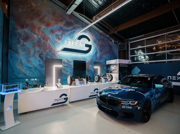 Ein recht dunkler Raum auf dem sehr viel blau zu erkennen ist. Rechts steht ein blauer BMW M5 links an der Wand hängt ein bleuchtetes Arnold NextG Logo und untendrunter befinden sich Tische mit Exponaten und ein Ferneher.