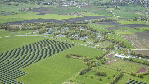 Eine Drohnenaufnahme des New Mobility Campus in Mengen/Hohentengen. Vorne Links sieht man mehrere Reihen von Solarfelder und rechts befindet sich ein Stadion. In der Mitte ist der Campus mit Hangar, ein altes Krankenhaus... zu sehen und im Hintergrund liegt die Stadt Hohentengen.