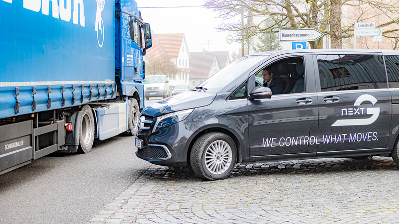 Links fährt ein blauer LKW auf der Straße vorbei. Von rechts kommt eine mit Arnold NextG Logos beklebte V-Klasse und blickt nach links. Auf dem Fahrersitz sitzt keiner, nur auf dem Beifahrersitz, sitzt ein Mann und schaut auf sein Handy.