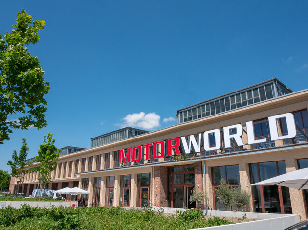 Standort München. Eingang der Motorworld München, einem langen eher flachen leicht rotem Gebäude, bei blauem Himmel und Sonnenschein.