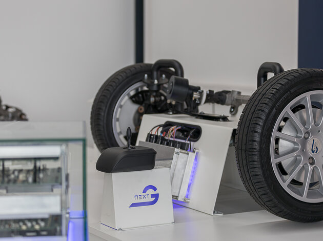 In der unteren Linken unteren Ecke sieht man unscharf, einen Anschnitt des NX NextMotion Exponates. Im Hintergrund befindet sich die Vorderachse, samt Rädern eines Autos, das verkabelt ist mit einem NX NextMotion Steuergeräts, welches man mit einem davor liegenden Joystick nach links und rechts bedienen kann.