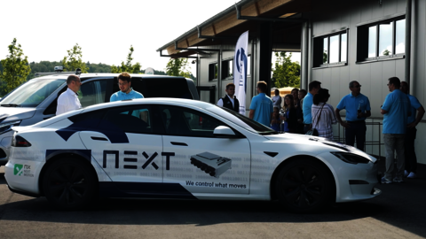 Ein weißer Tesla gebrandet mit NextG steht bei Sonnenschein vor einer Halle. Hinter dem Tesla steht ein weiteres Auto und mehrere Menschen.