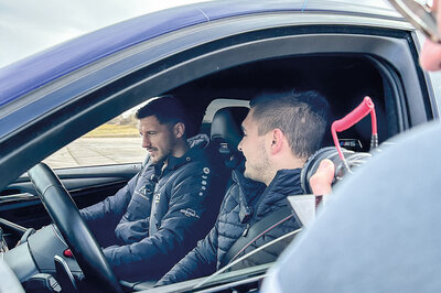 Zwei braunhaarige junge Männer sitzen in einem Auto und reden Miteinander. Eine weitere Person steht vor dem heruntergefahrenen Fahrerfenster und filmt in das Auto hinein.