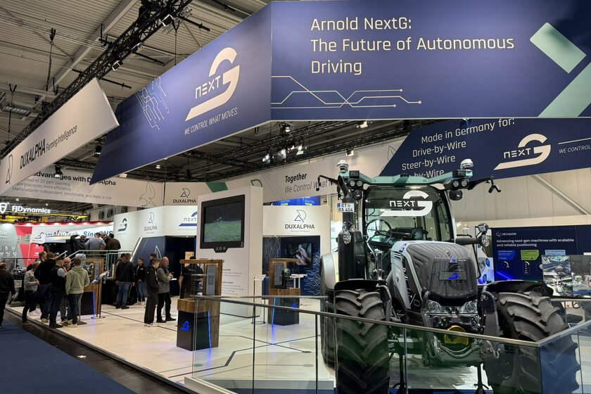 Am rechten Bildrand steht ein silberner Traktor mit großem NextG Logo auf der Frontscheibe. Der Traktor steht auf auf unserem Messestand der Agritechnica 2025, mit weißem Boden, Blauen Bannern mit Text an der Decke und im Hintergrund einen großen Fernseher.