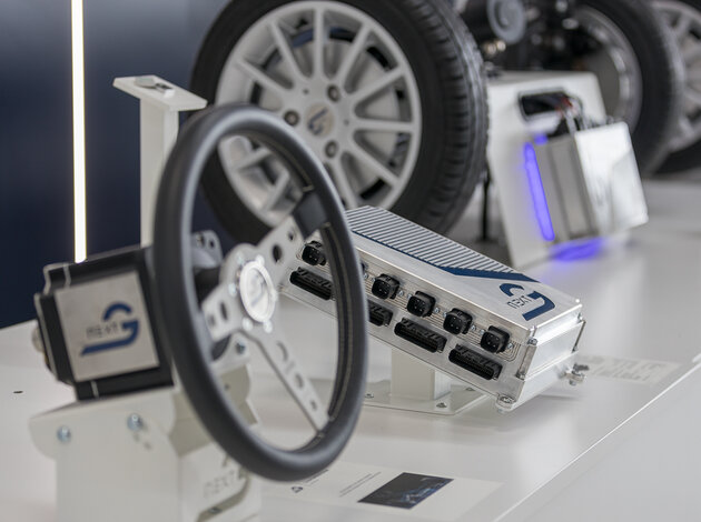 Auf einem Tisch befinden sich mehrere Objekte die schräg übers Bild verlaufen. Links unten befindet sich ein Lenkrad mit einem Motor hinten dran, in der Mitte liegt ein NX NextMotion Zentralsteuergerät und im Hintergrund befindet sich eine Platooning Anwendung von zwei Fahrzeugachsen die man mit einem Joystick lenken kann.