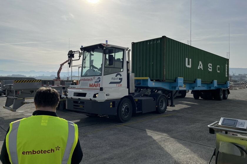 Ein weiß-grauer Terminal Truck mit grünem Container Auflieger hinten dran, welcher bei Sonnenschien auf einem Betonplatz steht.