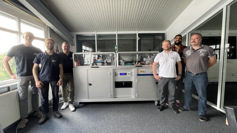 Jeweils 3 Personen stehen links und rechts neben einer Schulterhohen 2m langen Box, in einem Büro. 