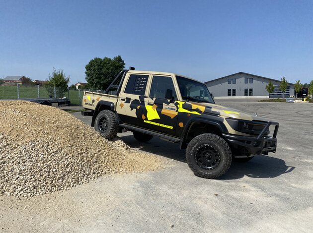 Modifizierter Toyota Landcruiser mit Tarnfolierung und Offroad-Reifen vor einem Schotterhaufen auf dem Testgelände.