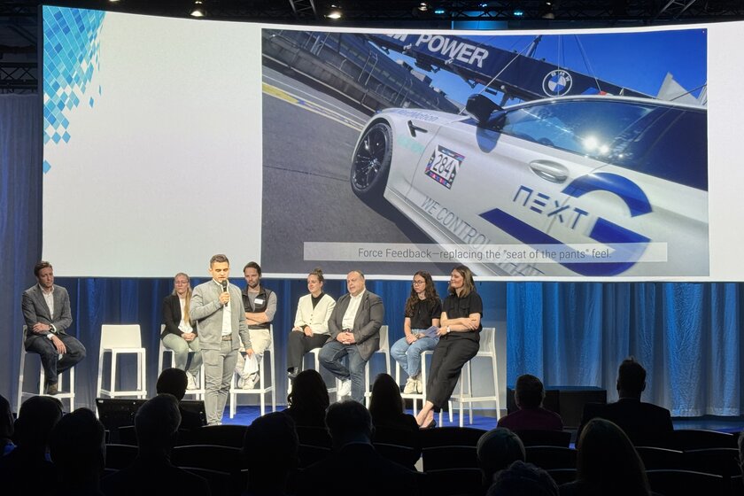 Vor einer sehr großen Leinwand auf der ein Arnold NextG Auto drauf ist, sitzen mehrere Personen auf einer Bühne. Vor ihnen steht ein junger Mann (Kevin Arnold) der in ein Mikro redet. 