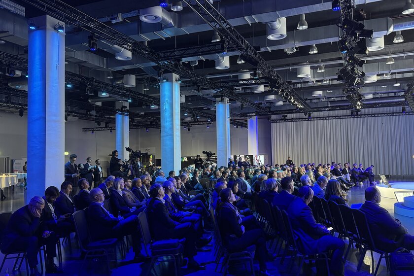 Viele Menschen sitzen in gestuhlten Reihen in einem großen Saal der blau beleuchtet ist.Viele Menschen sitzen in gestuhlten Reihen in einem großen Saal der blau beleuchtet ist.
