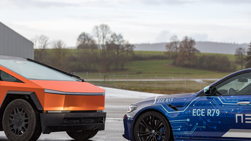 Zwei Autos stehen Schnauze an schnauze gegenüber. Das hintere ist ein orangener Cybertruck, das vordere ein blau gebrandeter BMW von Arnold NextG.