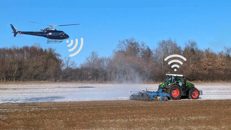 Auf einem Feld fährt ein Traktor mit Anbaugerät. Etwas hinter dem Traktor fliegt ein Helikopter, beide haben Wlan zeichen über sich.