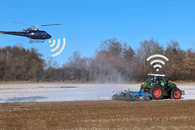 Auf einem Feld fährt ein Traktor mit Anbaugerät. Etwas hinter dem Traktor fliegt ein Helikopter, beide haben Wlan zeichen über sich.