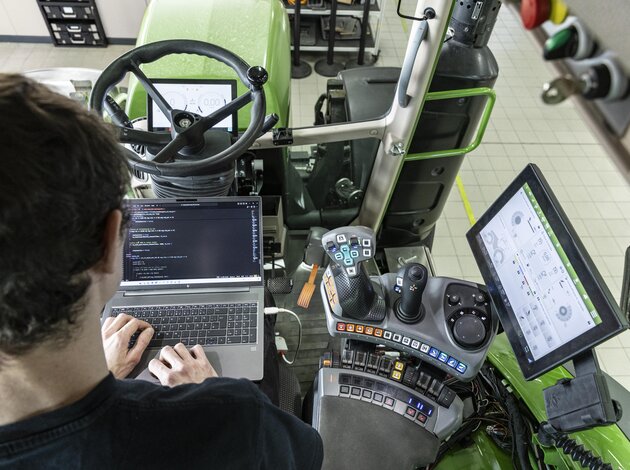 Einem Mitarbeiter der Firma Arnold NextG schaut man über die Schulter, durch das hintere Fenster einer Traktor Kabine, wärenddessen er an seinem Laptop, der auf seinem Schoß steht, an der Software arbeitet.