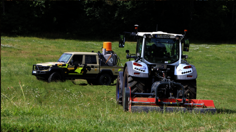 Ein Traktor mit Mähwerk fährt über eine Wiese, gefolgt von einem autonomen Land Cruiser mit Tarnlackierung und Tankeinheit auf der Ladefläche. Im Hintergrund ist ein Waldrand zu sehen. Die Szene zeigt den Einsatz moderner Drive-by-Wire-Technologie in der Landtechnik.