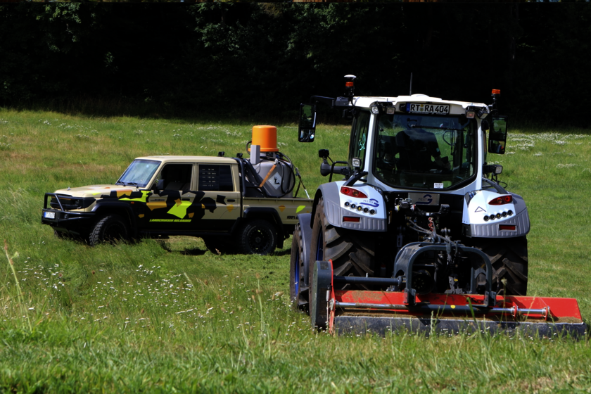 Ein Traktor mit Mähwerk fährt über eine Wiese, gefolgt von einem autonomen Land Cruiser mit Tarnlackierung und Tankeinheit auf der Ladefläche. Im Hintergrund ist ein Waldrand zu sehen. Die Szene zeigt den Einsatz moderner Drive-by-Wire-Technologie in der Landtechnik.