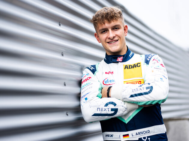 Ein blondhaariger lächelnder junger Mann, der Rennfahrer für das Team Arnold NextG, Mitgründer der Firma Arnold NextG, Luca Arnold lehnt sich mit der rechten Schulter gegen eine gewellte Wand im Arnold NextG weis blau türkisnen Rennanzug.