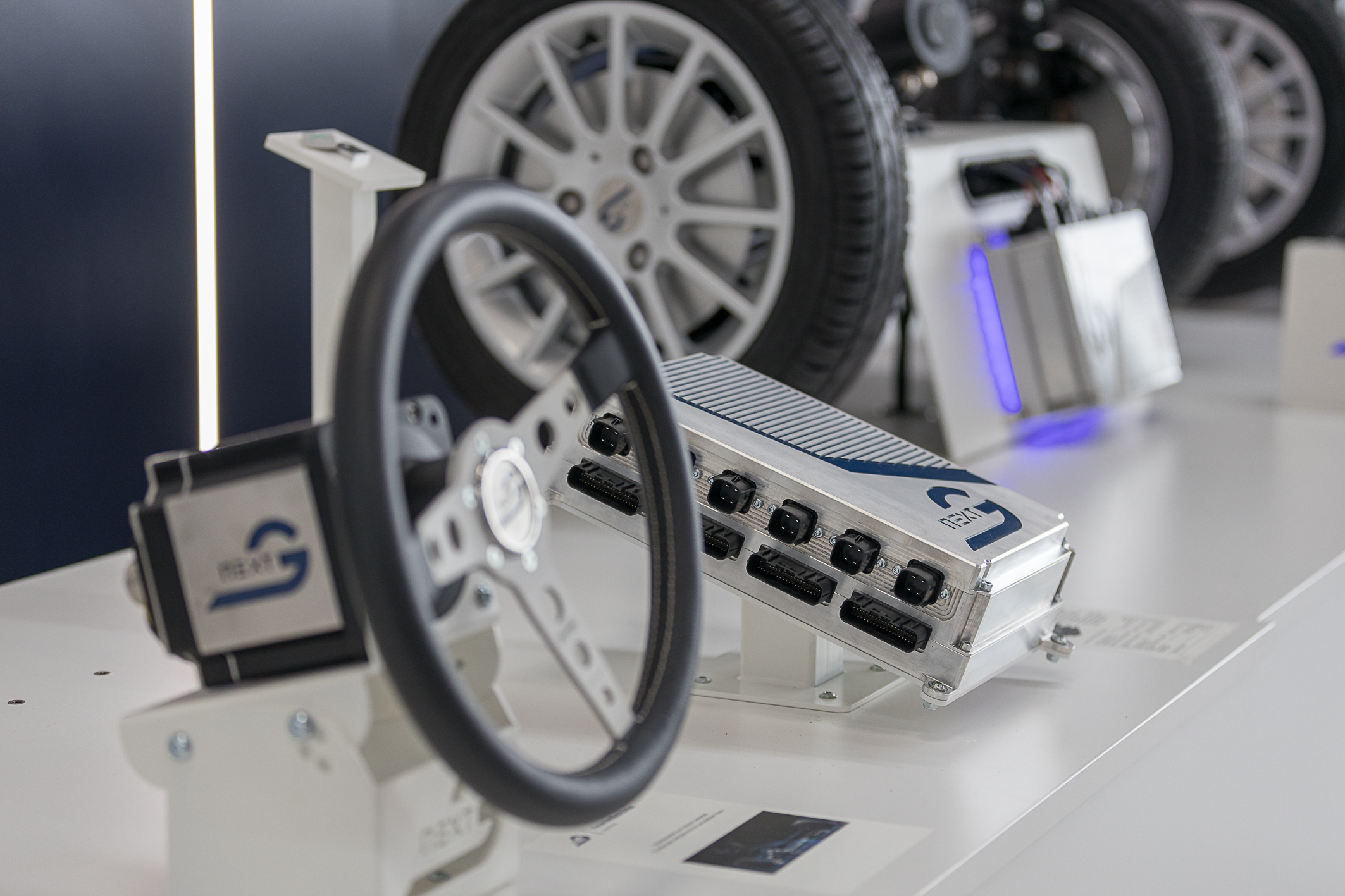 Auf einem Tisch befinden sich mehrere Objekte die schräg übers Bild verlaufen. Links unten befindet sich ein Lenkrad mit einem Motor hinten dran, in der Mitte liegt ein NX NextMotion Zentralsteuergerät und im Hintergrund befindet sich eine Platooning Anwendung von zwei Fahrzeugachsen die man mit einem Joystick lenken kann.