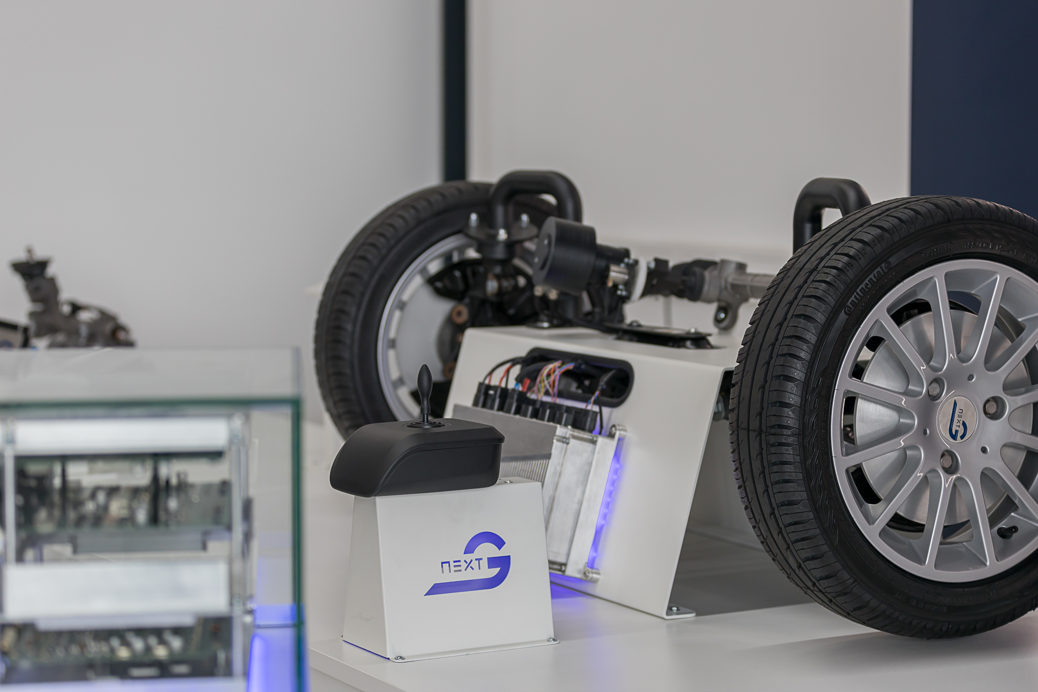 In der unteren Linken unteren Ecke sieht man unscharf, einen Anschnitt des NX NextMotion Exponates. Im Hintergrund befindet sich die Vorderachse, samt Rädern eines Autos, das verkabelt ist mit einem NX NextMotion Steuergeräts, welches man mit einem davor liegenden Joystick nach links und rechts bedienen kann.