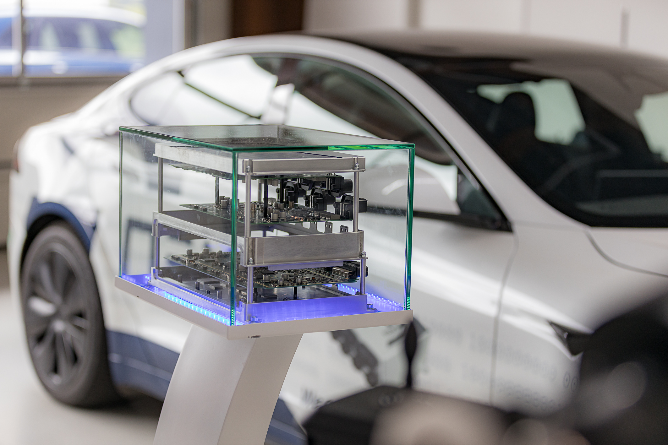 Im Vordergrund befindet sich das NX NextMotion, welches auseinandergezogen auf 3 Ebenen aufgebaut ist, sodass die Platinen im inneren zum Vorschein kommen. Das ganze wird in einer Glasvitrine präsentiert. Im Hintergrund steht unscharf ein weißer Tesla. 