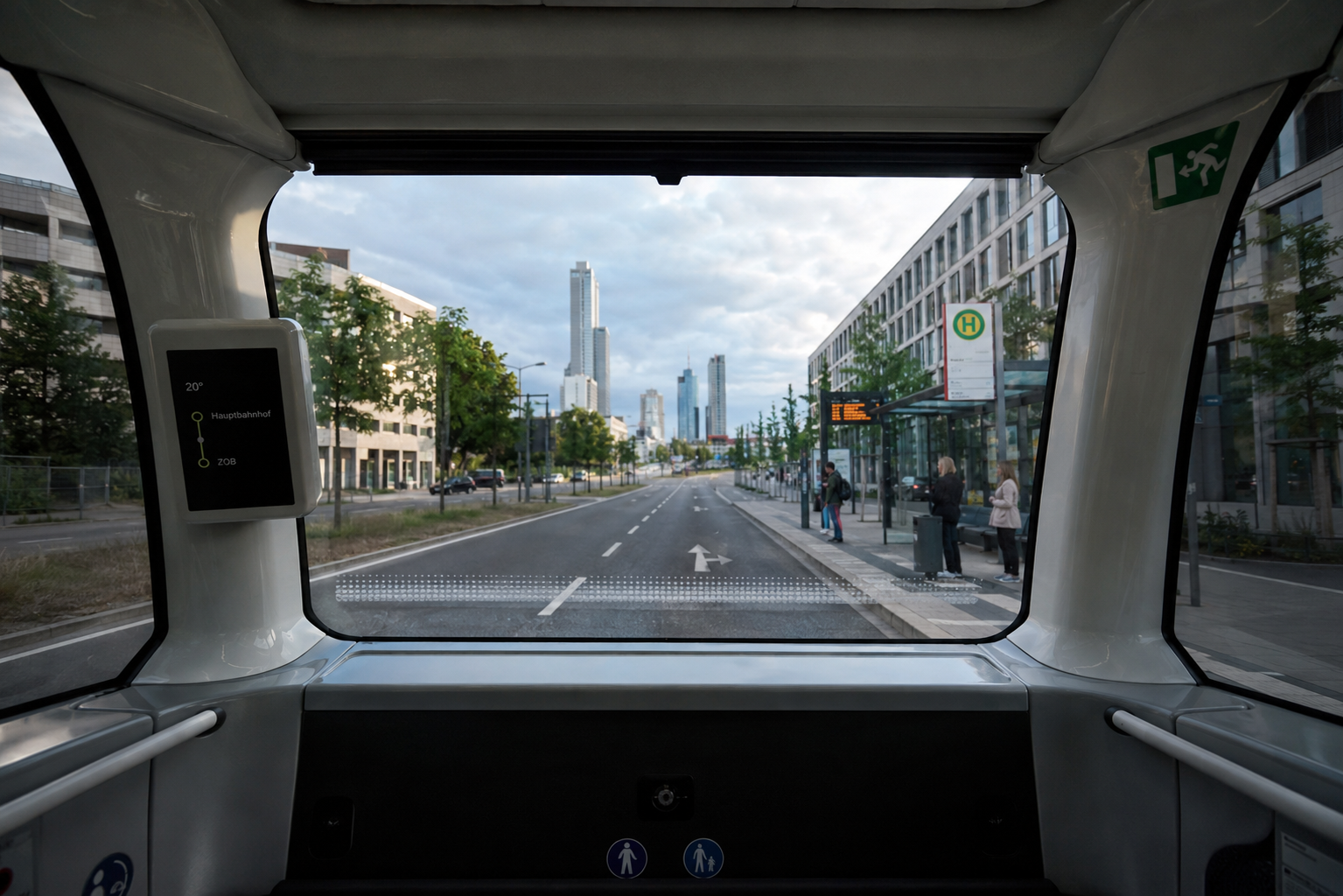 Der Blick aus der Frontscheibe eines autonomen Shuttle Bus, der auf der linken Fahrspur fährt. Rechts befindet sich eine Haltestellen, an der mehrere Personen schon warten. Im Hintergrund erkennt man eine Großstadt mit Hochhäuser.