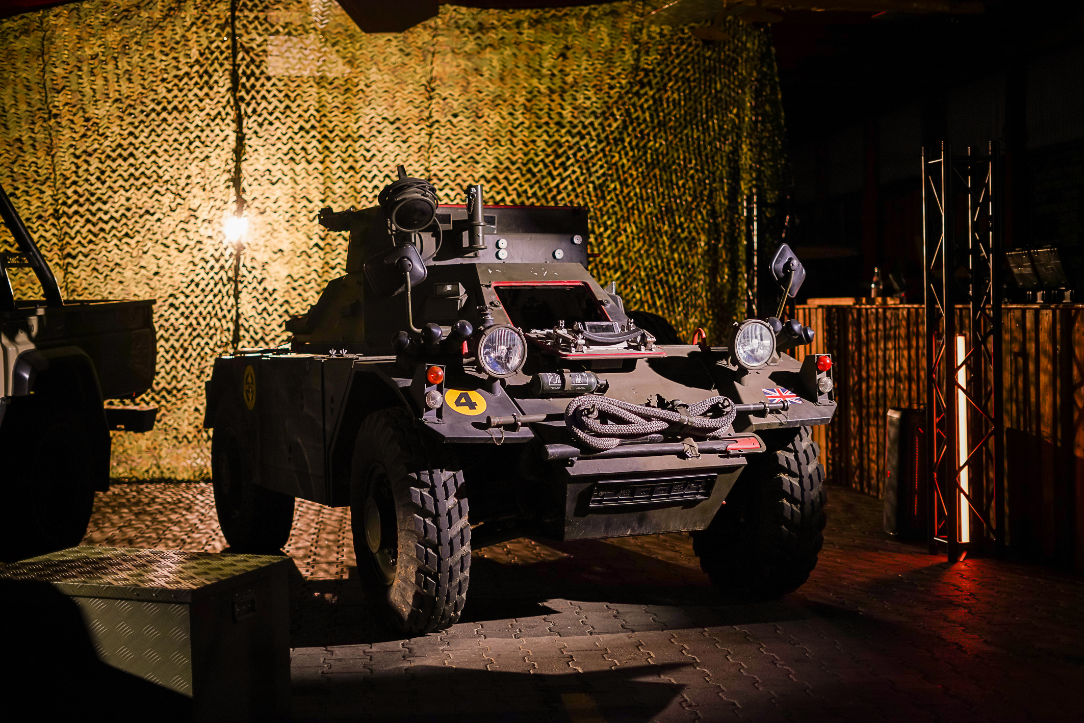 Ein älteres kleines Militärfahrzeug steht auf einem gepflasterten Steinboden mit Tarnnetz im Hintergrund. Das Bild ist in grün, gelblich dunkel gehalten 