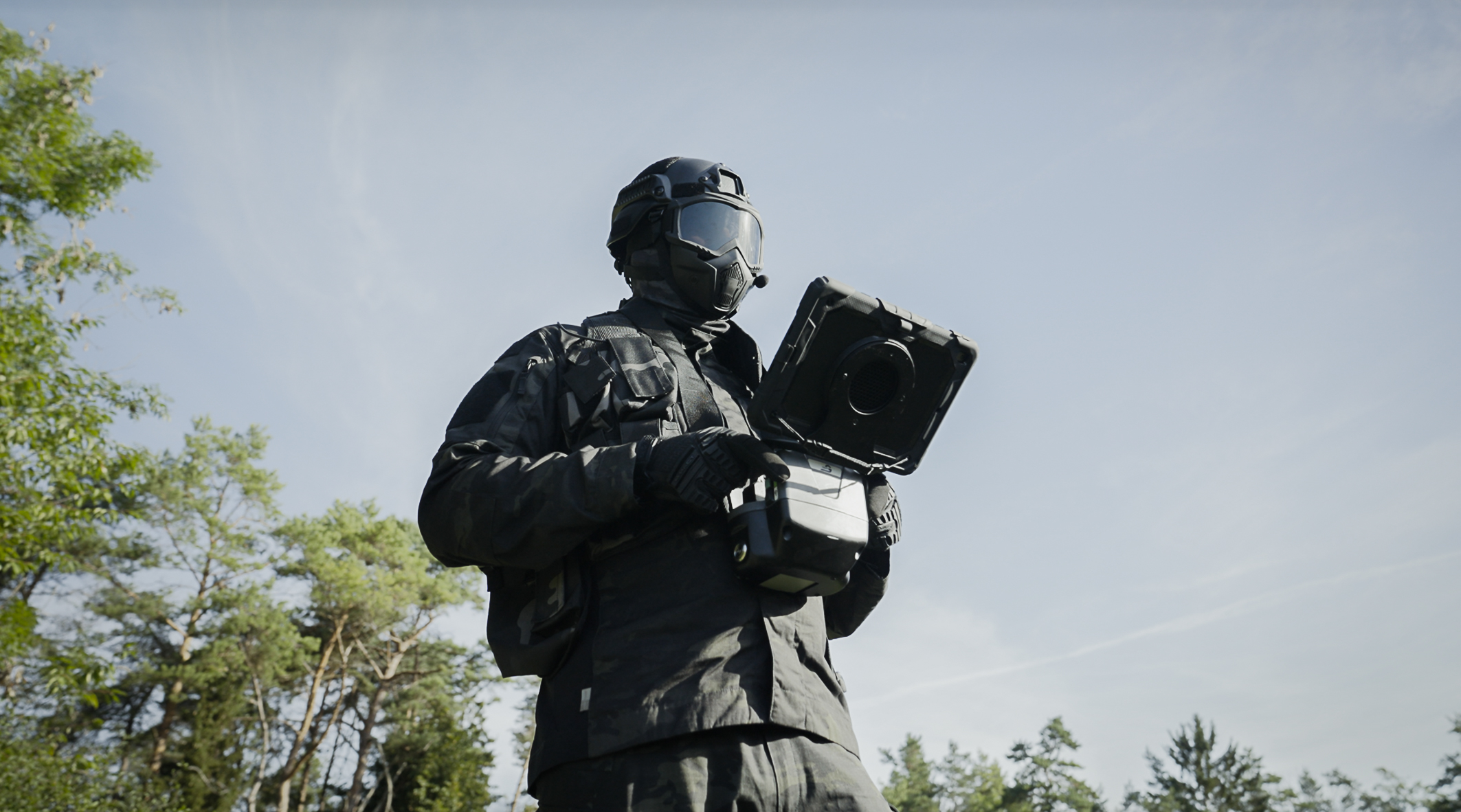 Eine Person steht bei blauem Himmel an einem Waldrand. Die Person ist in Tarnklamotten gehüllt und hat eine Gasmaske auf. In seinen Händen hält er eine Fernsteuerung mit IPad oben dran.