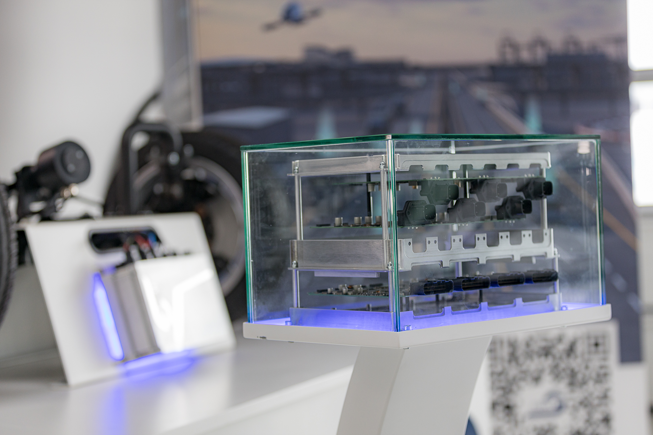 Hinter einem kleinen Glaskasten, welches ein Ausstellungsstück darstellt, befindet sich ein geöffnetes in Ebenen aufgebautes NX NextMotion. Im Hintergrund sieht man unscharf ein weitere Ausstellungsstück, eine Achse ebenfalls mit einem NX NextMotion.