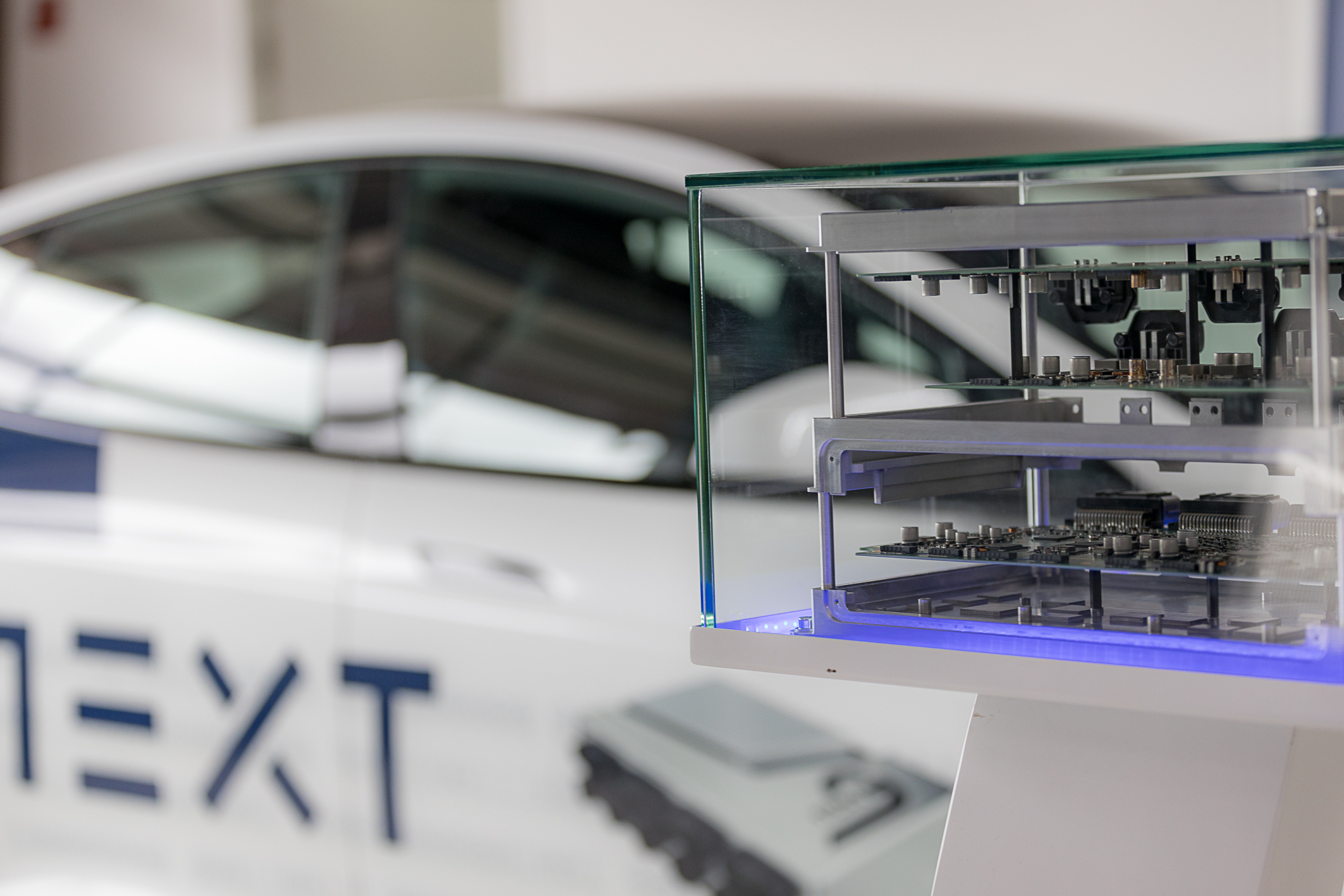 Hinter einem  Glaskasten, welches ein Ausstellungsstück darstellt, befindet sich ein geöffnetes in Ebenen aufgebautes NX NextMotion auf der rechten Seite. Im Hintergrund links sieht man unscharf einen weißen Tesla mit NextG Beklebung.
